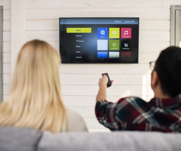 Young man with remote control choosing something to watch while sitting in front of tv set with his girlfriend near by